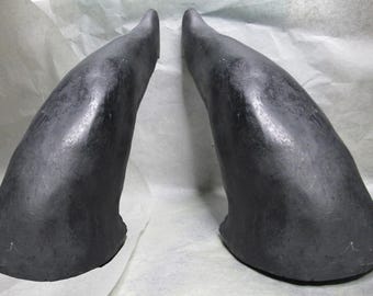 May include: Two black, curved, horn-shaped props made of a smooth, hard material. The horns are similar in size and shape, and they are both pointing upwards.
