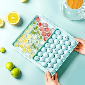 May include: Two light blue ice cube trays filled with round ice cubes. One tray contains clear ice cubes with fruit and herbs, the other has round blue ice cubes. Sliced lemons and green fruit are also visible.