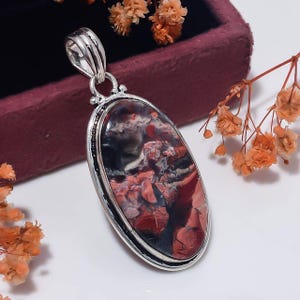 May include: An oval pendant featuring a polished, multi-coloured stone with deep red, black and white marbled patterns. The stone is set in a smooth, silver-toned metal frame with a decorative bail at the top for attachment to a necklace chain.