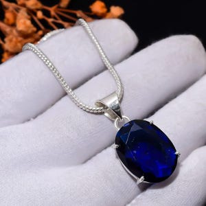 Può includere: Una collana in argento con un ciondolo in pietra preziosa blu scuro di forma ovale e sfaccettata. Il ciondolo è fissato in una semplice montatura in metallo a quattro griffe, sospeso a una sottile catena d'argento testurizzata su tessuto bianco.