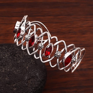 May include: A silver cuff bracelet featuring a repeating pattern of open, geometric shapes. The bracelet is set with oval, deep red gemstones and small, floral details. The bracelet is displayed on a dark, textured surface.