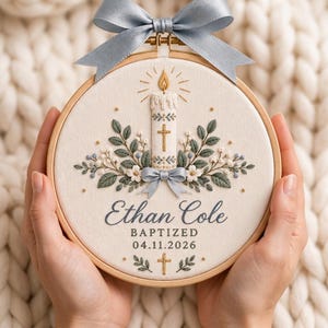Puede incluir: Un bastidor de bordado circular de madera con tela blanca que presenta un diseño bordado de una vela encendida, una cruz dorada y follaje verde. El texto dice Ethan Cole, BAPTIZED, 04.11.2026. Un lazo de cinta azul claro está sujeto arriba.