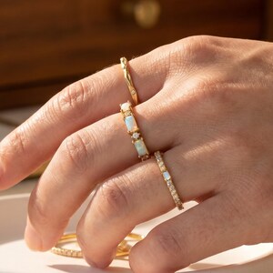 May include: A hand wearing several gold rings. The rings feature various designs, including a textured band with a small diamond, opal stones, and a band with small clear stones. The rings are displayed in natural light.
