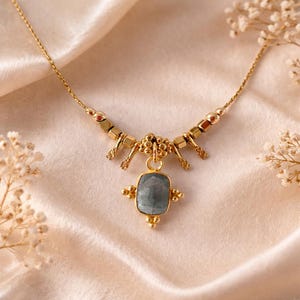 May include: A gold-toned necklace with a rectangular, gray-blue gemstone pendant. The necklace features a delicate chain and decorative gold elements. The pendant is adorned with small, gold embellishments, creating an elegant and sophisticated look.