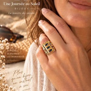 May include: A gold-toned rectangular ring featuring a textured surface and three dark, oval-shaped stones set vertically in the center. The ring is worn on a finger, showcasing its detailed metalwork and the contrast between the gold and dark stones.