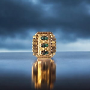 Könnte beinhalten: Ein goldfarbener Ring mit drei smaragdgrünen Edelsteinen, die in einem rechteckigen Design gefasst sind. Der Ring hat ein strukturiertes Band und wird auf einer reflektierenden Oberfläche präsentiert.