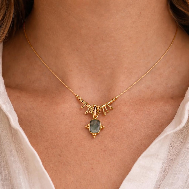 May include: A gold-colored necklace with a rectangular, light blue gemstone pendant. The necklace has intricate detailing around the pendant and along the chain, creating an elegant and delicate design. The necklace is worn against a white shirt.