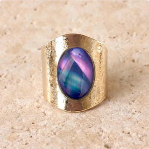 Könnte beinhalten: Ein breiter goldfarbener Ring mit einem gehämmerten, strukturierten Metallband und einem großen, ovalen Cabochon-Stein. Der Stein zeigt ein lebhaftes, irisierendes Farbspiel in tiefem Blau, Violett und Magenta mit einer glatten Oberfläche.
