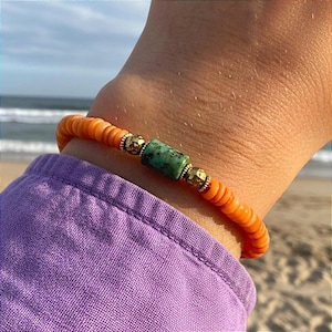 Könnte beinhalten: Ein elastisches Armband mit kleinen, runden, leuchtend orangefarbenen Scheibenperlen. In der Mitte befindet sich eine zylindrische Türkisperle mit dunkler Maserung, die von zwei kleinen, strukturierten, goldfarbenen Metallperlen flankiert wird.