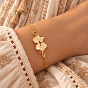 Op de afbeelding: Een goudkleurige armband met twee symmetrische ginkgo-bladbedels die in het midden met elkaar zijn verbonden. De armband is gemaakt met een fijn schakelkettingontwerp en heeft een gepolijste metalen afwerking die het licht reflecteert.