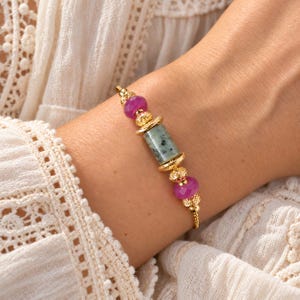 Könnte beinhalten: Ein Armband mit goldener Kette, das in der Mitte eine zylindrische, grün gesprenkelte Steinperle aufweist, flankiert von zwei goldenen Filigran-Abstandshaltern, gefolgt von zwei facettierten magentafarbenen Perlen und weiteren goldenen Akzenten.