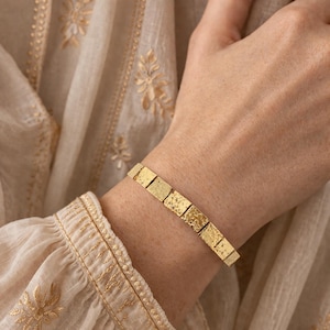 May include: A gold bracelet featuring a series of square blocks. The bracelet is worn on a wrist, with a cream-coloured blouse in the background. The bracelet's texture appears to be slightly rough.