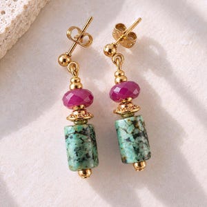 May include: A pair of gold-tone drop earrings featuring a faceted magenta bead, a small gold-tone spacer, and a cylindrical turquoise-colored stone with dark veining. The earrings are finished with gold-tone metal posts and butterfly backs.