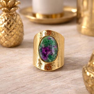 Peut inclure: Une bague dorée avec une pierre ovale verte et violette. La bague a un large anneau texturé. Décorations dorées en forme d'ananas et bougeoir en arrière-plan. La pierre de la bague mesure environ 2 cm de long.