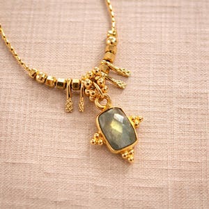 May include: A gold-toned necklace with a rectangular, light green gemstone pendant. The necklace features a delicate chain and decorative elements, including small beads and ornate accents. The pendant is framed by a gold setting with additional gold embellishments.