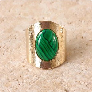 Pode incluir: Um anel largo em tom dourado com uma pedra preciosa de malaquita verde de formato oval ao centro, com padrões de faixas escuras. A banda de metal tem um acabamento martelado texturizado que contrasta com a superfície lisa e polida da pedra.