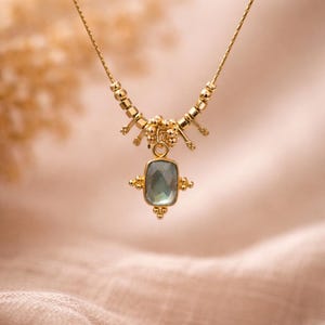 May include: A gold-colored necklace with a rectangular, light-blue gemstone pendant. The necklace features a delicate chain and decorative gold beads. The pendant is framed by small gold accents, creating an elegant and sophisticated look.