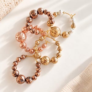 Op de afbeelding: Drie oorringen met getextureerde, poreuze kralen in metallic tinten koper, brons en goud. De cirkelvormige ringen zijn versierd met bolvormige kralen van verschillende groottes, waaronder enkele met een glanzende afwerking en andere met een matte, vulkanische textuur, gerangschikt langs dunne metalen draden.