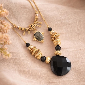 May include: Two gold necklaces with black and gold beads and pendants. One necklace features a large, faceted black stone pendant. The other has a smaller, rectangular stone pendant and a decorative gold charm. The necklaces are displayed on a beige fabric.