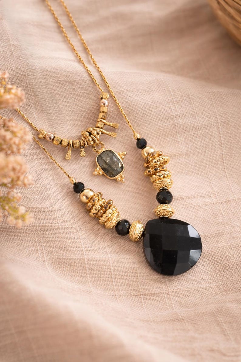 May include: Two gold necklaces with black and gold beads and pendants. One necklace features a large, faceted black stone pendant. The other has a smaller, rectangular stone pendant and a decorative gold charm. The necklaces are displayed on a beige fabric.