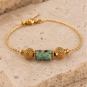 Peut inclure: Bracelet doré avec une perle cylindrique turquoise et deux perles rondes texturées. Le bracelet a une chaîne délicate et un fermoir décoratif. Le bijou est présenté sur une surface de ton neutre.