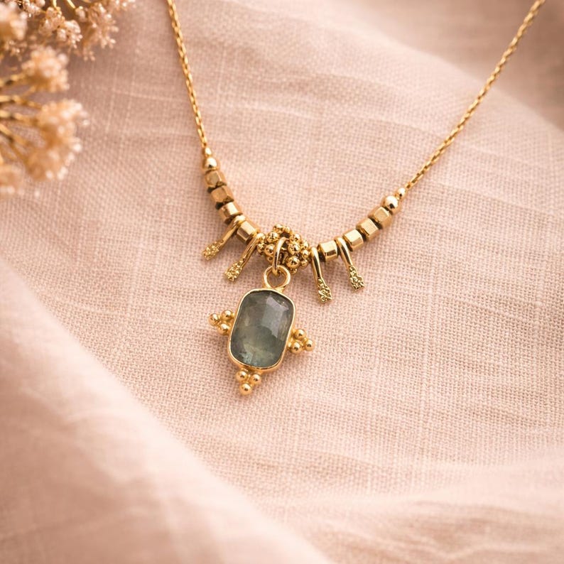 May include: A gold-colored necklace with a rectangular, light green gemstone pendant. The necklace features a delicate chain and decorative elements. The pendant is framed by a gold setting with small, round accents. The necklace is displayed on a light pink fabric.