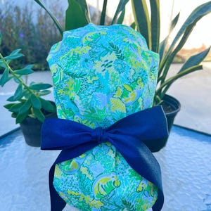 May include: A patterned fabric item in shades of green, blue, and yellow, tied with a navy blue bow. The fabric features a repeating design of stylized sea creatures and floral elements. The item is likely a gift or decorative item.