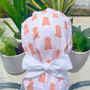 May include: A white scrub cap featuring a repeating pattern of peach-colored dog illustrations and pink bows. The cap is tied with a large white bow at the back. The cap is designed for medical professionals or those in the healthcare field.