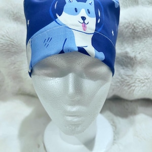 May include: A navy blue surgical scrub cap featuring a cartoon dog in a space helmet. The dog is light blue and white with a pink tongue. White stars are scattered across the cap's background.