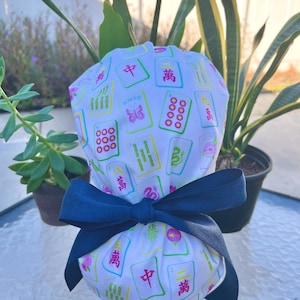May include: A white scrub cap featuring a colorful Mahjong tile pattern with pink, green, and yellow accents. The cap is tied with a navy blue bow. The cap is designed for medical professionals.
