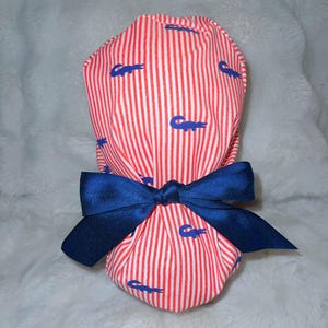 May include: A fabric item, possibly a hat or gift wrap, featuring a red and white striped pattern with blue crocodile motifs. A navy blue bow is tied around the item. The item is on a white, fluffy surface.