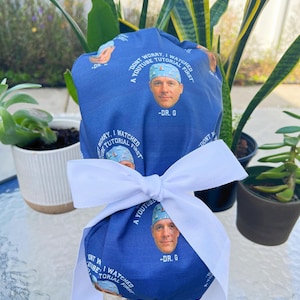 Puede incluir: Un gorro quirúrgico azul con el texto "DON'T WORRY, I WATCHED A YOUTUBE TUTORIAL FIRST" y una foto de un hombre. El gorro está atado con una cinta blanca. Hay plantas en el fondo.