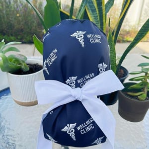 May include: Navy blue scrub cap with a white bow. The cap features a pattern of white caduceus symbols and the words "Wellness Clinic". It is tied with a white ribbon.