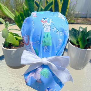 May include: A light blue surgical scrub cap with a white bow. The cap features a repeating pattern of Dalmatian dogs in hula skirts, palm trees, and other tropical elements. The bow is tied at the back.