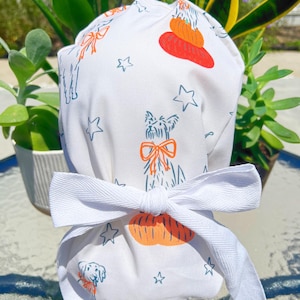 May include: White fabric scrub cap with a repeating pattern of dogs, pumpkins, and stars in orange and blue. The cap is tied with a white ribbon bow. The cap is designed for medical professionals.