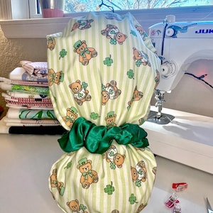 May include: A green and white striped fabric item with a pattern of teddy bears wearing green hats and shamrocks. The item is tied with a green satin scrunchie. Sewing supplies and a sewing machine are in the background.