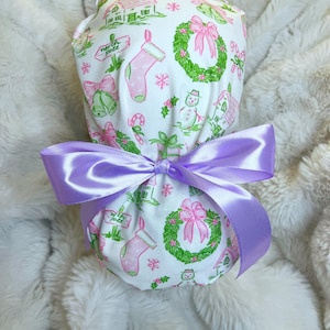 May include: A white scrub cap featuring a Christmas pattern with green houses, pink stockings, and wreaths. It is tied with a lavender satin bow. The design is festive and cheerful, perfect for the holiday season.