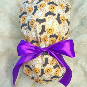 Puede incluir: Un gorro quirúrgico con temática de Halloween, con calabazas naranjas, fantasmas blancos y murciélagos negros sobre un fondo crema. Está atado con un lazo de satén morado brillante.