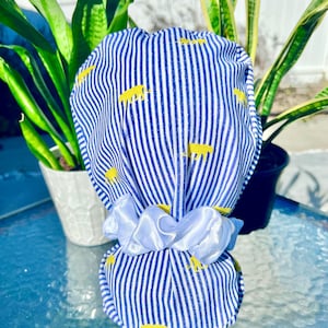 May include: A blue and white striped fabric item with yellow animal shapes, tied with a white satin ribbon. The fabric has vertical stripes. The item is likely a hair accessory or a gift bag.