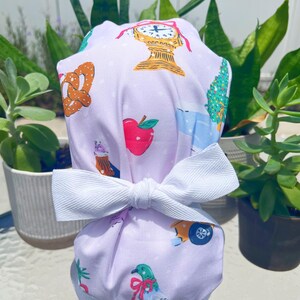 May include: A lilac scrub cap with a white bow. The cap features a pattern of illustrations, including a clock, apple, pretzel, and a car. The bow is tied at the front of the cap.