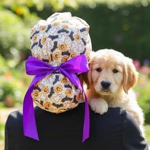 Puede incluir: Un gorro quirúrgico con temática de Halloween con un lazo morado, usado por una persona que sostiene un cachorro golden retriever. El gorro presenta un patrón de calabazas, fantasmas y murciélagos sobre un fondo crema.
