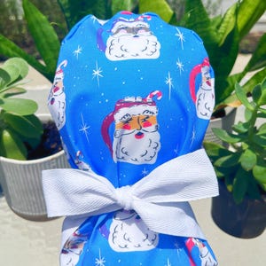 May include: A blue scrub cap with a white bow. The cap features a repeating pattern of Santa Claus mugs with candy canes and snowflakes. The Santa mugs have a red and white design. The bow is made of white fabric.
