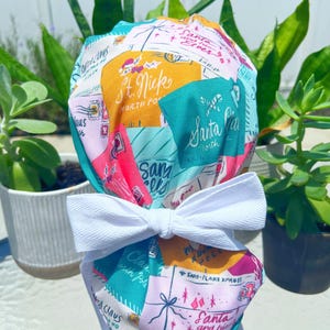 May include: A festive scrub cap with a white bow, featuring a patchwork design. The cap displays Christmas-themed text and illustrations of Santa Claus, elves, and North Pole imagery in pink, teal, yellow, and white.