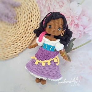 Op de afbeelding: Gehaakte pop met donkere huid, zwart krullend haar en gouden oorbellen. De pop draagt een witte top, een paarse rok met gele details en een blauw en turquoise lijfje. De pop heeft een roze hoofddoek.