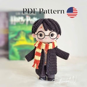 以下が含まれることがあります： 黒い髪、丸いメガネ、赤と黄色のストライプのスカーフをしたキャラクターの編み人形。人形は黒いコートと灰色のズボンを着ています。画像には「PDFパターン」というテキストと小さなアメリカ国旗が含まれています。