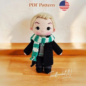 May include: A crocheted doll with blonde hair, wearing a black coat and a green and white scarf. The doll has a gray shirt and black trousers. The text "PDF Pattern" and a US flag are in the background. The doll is standing on a wooden surface.