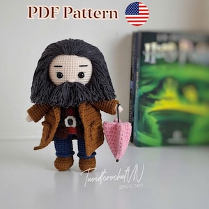 Puede incluir: Muñeco de ganchillo de un personaje con barba larga, que lleva un abrigo marrón, pantalones azules y sostiene un paraguas rosa. El muñeco tiene barba y pelo negros. El texto "PDF Pattern" y la bandera americana son visibles.