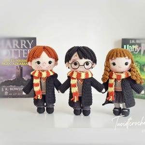 May include: Three crocheted dolls inspired by the Harry Potter series, each with distinct hair colors and round glasses. They wear black robes and striped scarves, standing in front of book covers. The dolls are handmade and feature intricate details.