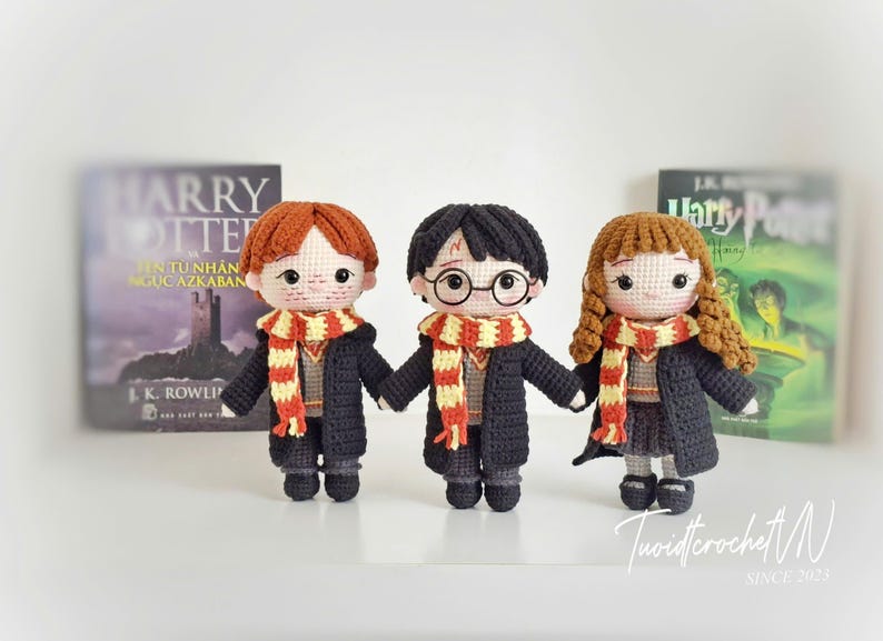 May include: Three crocheted dolls inspired by the Harry Potter series, each with distinct hair colors and round glasses. They wear black robes and striped scarves, standing in front of book covers. The dolls are handmade and feature intricate details.