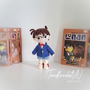 May include: A handmade crochet doll of a character wearing a blue jacket, shorts, and red bow tie, with glasses. The doll is standing in front of a book with the word "Conan" on the cover. The doll is made with brown, blue, red, white, and beige yarn.
